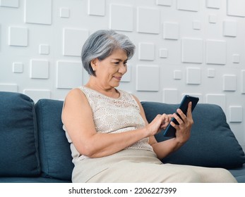 Happy Asian Senior Woman Sit On Couch, Holding And Looking At Digital Tablet Screen In Hand In Living Room. Elderly Female Using Smart Device, Computer At Home. Older People Use Technology With Easy.