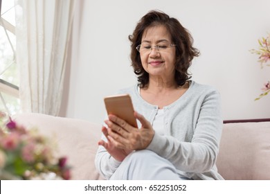 Happy Asian Senior Woman Sitting On Sofa And Using A Smart Phone