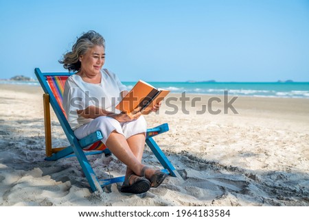 Happy Asian senior woman sitting on beach chair on the beach with reading a book. Retirement elderly female resting on sunbed by the sea relax and enjoy outdoor lifestyle activity in summer vacation