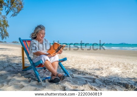 Happy Asian senior woman travel on summer beach holiday vacation. Elderly retired female relax and enjoy outdoor lifestyle resting on beach chair with reading a book at the sea in summer sunny day.