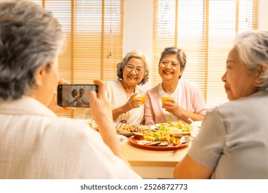 Happy Asian senior women friends having dinner together at home. Elderly retired woman enjoy indoor lifestyle using mobile phone taking picture during eating healthy food together on dining table. - Powered by Shutterstock