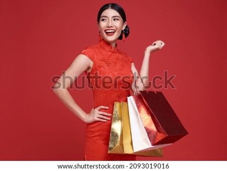 Happy Asian shopaholic woman wearing traditional cheongsam qipao dress holding shopping bag isolated on red background. Happy Chinese new year	