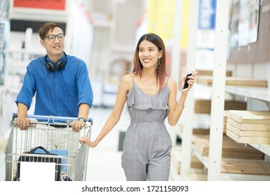 Happy Asian Shopper Couple With Shopping Cart Walk In Ales Of Furniture Card Box Shelf In Hypermarket.