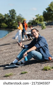 Happy Asian Skater With Soda Can And Mobile Phone Sitting On Embankment Near Blurred Friends