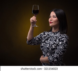 Happy Asian Sommelier Woman Holsing A Glass Of Red Wine In Front Of Her. Lady With Black Hair Looking At Wine's Individual Colour Isolated On Dark.