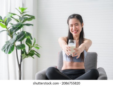 Happy Asian Sports Girl In Sportswear Sitting On Chair And Drinking Pure Milk From Glass After Exercise In Home. Healthy Woman Smiling And Drink Milkshake Protein. Healthcare And Weight Loss Concept.