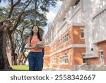 Happy asian student walking on university campus holding books and backpack, looking away smiling, with trees and school building in background