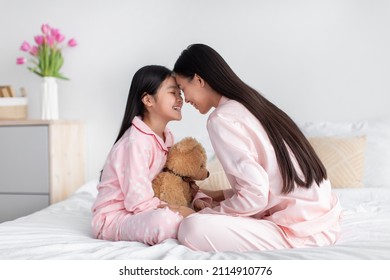 Happy Asian Teen Girl With Toy And Millennial Woman In Pink Pajamas Enjoy Time Together Sitting On Bed In Bedroom Interior, Profile. Family Relationship, Love, Mother And Daughter Together At Home