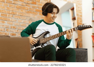 Happy Asian Teen Guy Playing Electric Guitar And Using Laptop Computer Recording New Song Sitting At Home, Wearing Wireless Headphones. Musical Instruments And Hobby Concept. Selective Focus - Powered by Shutterstock