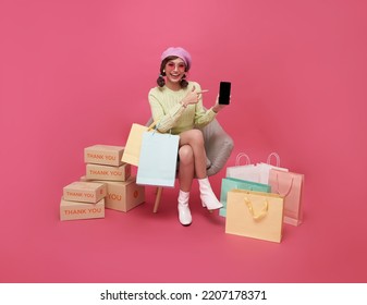 Happy Asian teen woman sitting on sofa holding shopping bags and smartphone isolated on pink background, Shopper or shopaholic concept.