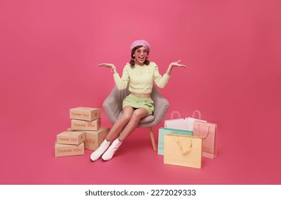 Happy Asian teen woman sitting on sofa with shopping bags and gift box isolated on pink background, Shopper or shopaholic concept.