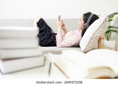 Happy asian teenage girl wearing headphones lying on the couch,resting and listening to music online on mobile phone,enjoy and relax reducing tension after reading,studying textbook,relaxation concept