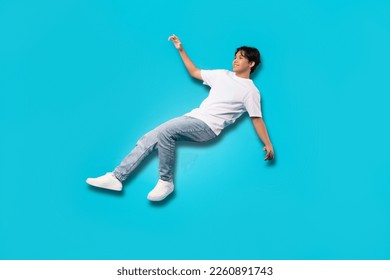 Happy Asian Teenager Boy Falling Posing In Mid Air Smiling Looking Aside Over Blue Background. Studio Shot Of Positive Teen Guy Experiencing Zero Gravity. Full Length - Shutterstock ID 2260891743