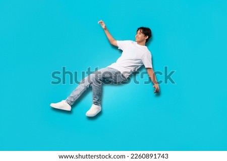 Happy Asian Teenager Boy Falling Posing In Mid Air Smiling Looking Aside Over Blue Background. Studio Shot Of Positive Teen Guy Experiencing Zero Gravity. Full Length
