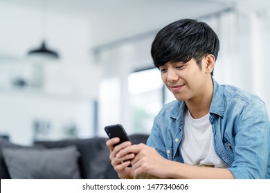 Happy asian teenager using smart phone and smiling on sofa living room at home. Asian man holding and using cellphone for searching data and social media on internet. Teenager and Technology concept.
