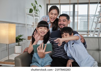 Happy Asian Thai Parents, And Children Online Video Call With Family Via Internet On Sofa In Home Living Room, Lovely Weekend, And Domestic Wellbeing Lifestyle With Internet Technology Mobile Phone.