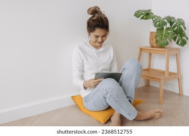 Happy asian Thai woman bun hair, sitting on floor leaning on wall, using tablet, working or watching series at apartment in winter day. - Powered by Shutterstock