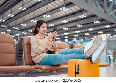 Happy Asian Tourist Woman Using Mobile Phone And Put Leg On Suitcase Traveling Between Waits For Flight In Airport Terminal, Browse Internet, Flight Check In, Tourist Journey Trip Concept