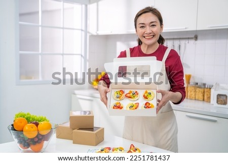 Happy Asian woman bakery shop owner holding fruit tart in delivery box for customer order. Bakery chef baking pastry and cake in the kitchen. Small business entrepreneur and food delivery concept.