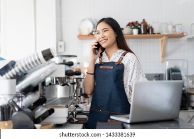 Happy Asian Woman Barista Working On Laptop Smiling And Talking On Smartphone, Accepting Writing Order At A Coffee Shop. Waitress In Cafeteria Small Business Owner, Online Purchase And Service Concept