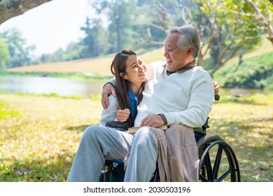 Happy Asian Woman Caring And Hugging Senior Man Grandfather Sitting On Wheelchair In The Park. Elderly Retired Male Relax And Enjoy Outdoor Leisure Activity With Daughter. Family Relationship Concept.