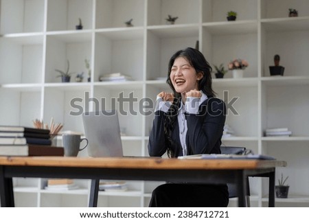 Happy Asian woman celebrates victory while receiving good news while working in modern office. Excited about success.