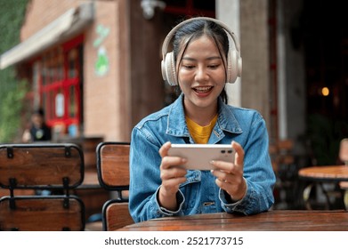 A happy Asian woman in a denim jacket wearing headphones is enjoying playing a mobile game or watching a video on her phone while sitting at an outdoor table of a restaurant.