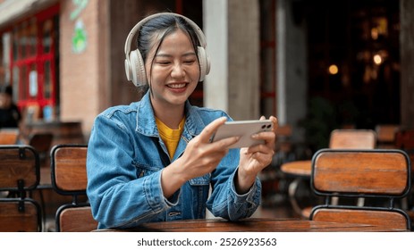 A happy Asian woman in a denim jacket wearing headphones is enjoying playing a mobile game or watching a video on her phone while sitting at an outdoor table of a restaurant.