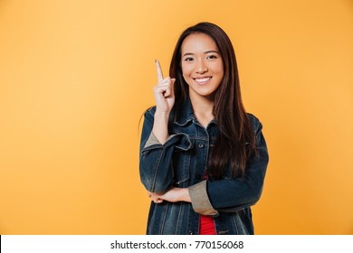 Happy Asian Woman In Denim Jacket Pointing Up And Looking At The Camera Over Yellow Background