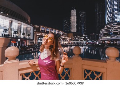 Happy Asian Woman In Dress Raising Hands In Front Of The Skyscrapers In Night City Of Dubai, UAE. Travel And Tourist Destinations