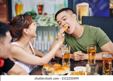 Happy Asian Woman Feeding Her Boyfriend With Fried Chicken While They Drinking Beer In Cafe Together With Their Friends