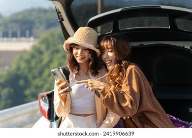 Happy Asian woman friends enjoy and fun outdoor lifestyle road trip countryside on summer holiday vacation. Two attractive girl sitting on car trunk using mobile phone taking selfie together. - Powered by Shutterstock