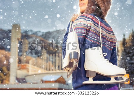 Happy asian woman going to ice skating outdoor. She dressed in pullover and warm hat. Holding skates shoes. Healthy lifestyle and sport concept at olympic stadium. Winter weather with snowy blizzard