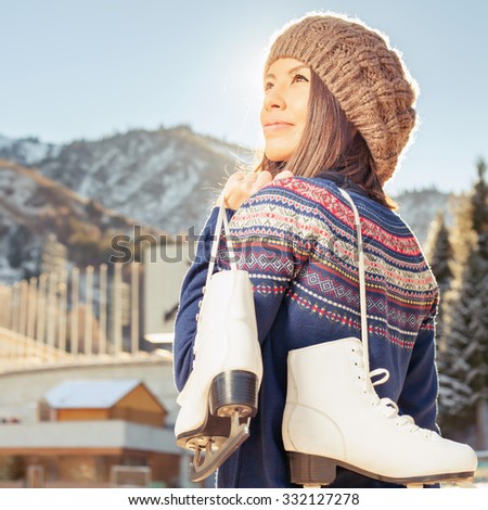 Happy asian woman going to ice skating outdoor. She dressed in pullover and warm hat. Holding skates shoes. Healthy lifestyle and sport concept at olympic stadium, beautiful nature. Mountain landscape
