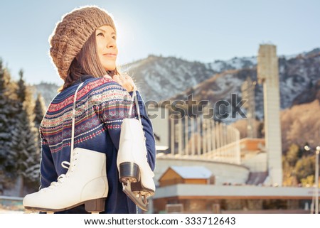 Happy asian woman going to ice skating outdoor. She dressed in pullover and warm hat. Holding skates shoes. Healthy lifestyle and sport concept at olympic stadium, beautiful nature. Mountain landscape