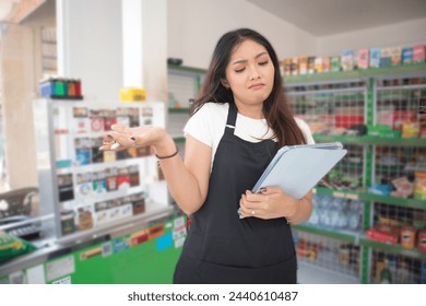 Happy Asian woman as groceries or modren market staff checking products and goods on the display shelf while holding tablet and wearing black apron