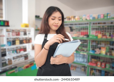 Happy Asian woman as groceries or modren market staff checking products and goods on the display shelf while holding tablet and wearing black apron