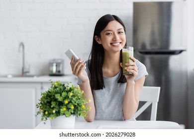 Happy Asian Woman Holding Cellphone And Glass Of Homemade Smoothie Near Blurred Potted Plant