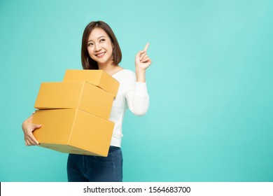 Happy Asian Woman Holding Package Parcel Box And Pointing Finger To Empty Copy Space Isolated On Light Green Background, Delivery Courier And Shipping Service Concept