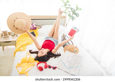 Happy Asian Woman Lying Down On Bed Showing Passport And Plane Ticket With Many Items To Preparing To The Beach Trip On Weekend. Young Pretty Girl Planning To Travel On Holiday At Home.