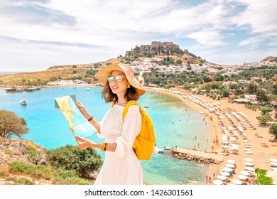 Happy asian woman with map and backpack wearing big vacation hat at the background of paradise view to Lindos town and beach on a Rhodes Island, Greece - Powered by Shutterstock