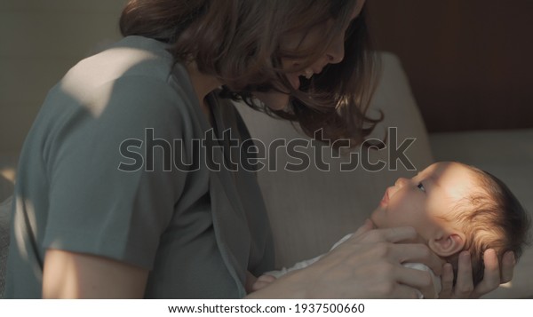 happy asian woman\
mother sitting on chair and holding sleepy baby infant in arm.\
female touching kid\'s hair and head lull to calm crying baby down\
and sleep. natural\
authentic.
