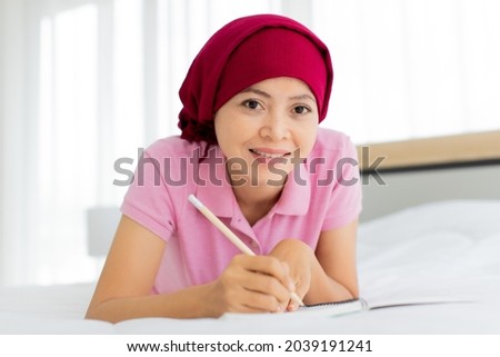 Happy Asian woman on pink shirt and cover head by red scarf as breast cancer patient in hospital smiling while lying down on clinical therapy bed and enjoy note jot on diary by pencil on bright day