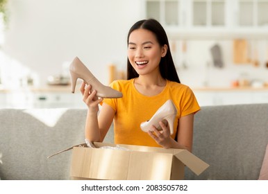 Happy Asian Woman Received Package, Unpacking Cardboard Box, Holding And Looking At High Heel Shoes, Sitting On Sofa At Home. Excited Female Buyer Satisfied With Online Shopping