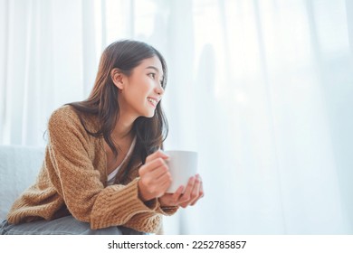 Happy asian woman relaxing drinking hot coffee or tea in holiday morning vacation on armchair at home, Cosy scene, Smiling pretty woman drinking hot tea in autumn winter. copy space. - Powered by Shutterstock