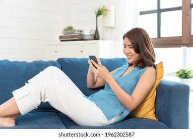 Happy Asian Woman Relaxing On Comfortable Sofa Using Smartphone Chatting In Social Networks, Watching Funny Videos At Home.