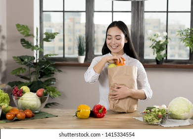 Happy Asian Woman Removing Vegetables From Grocery Bag On Wooden Kitchen Counter