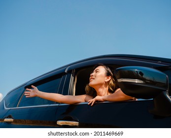 Happy Asian woman with short hair raising a hand against the wind while traveling by car. Attractive female travelers enjoy and smiling at the view outside the car, on the local road, summertime.