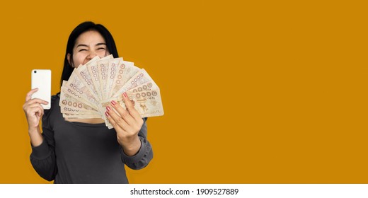 Happy Asian Woman Showing Thai Baht Money Another Hand Holding Smart Phone Over Orange Banner Background 