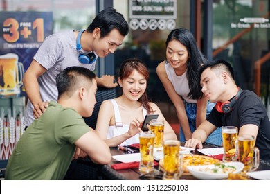 Happy Asian Woman Sitting With Mobile Phone And Showing To Her Friends Funny Photos On It During Their Meeting In Cafe
